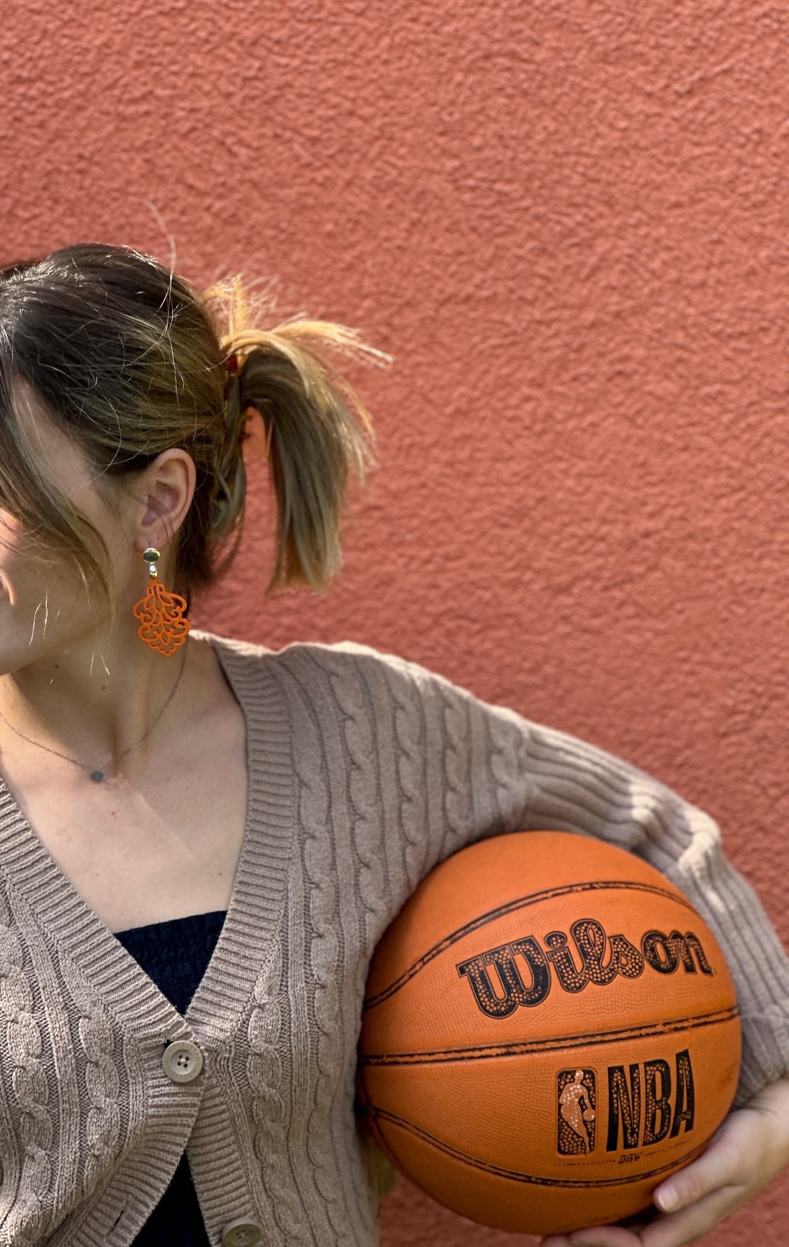 ine Frau mit blonden Haaren, die zu einem Pferdeschwanz gebunden sind, trägt einen orangefarbenen Ohrring mit kunstvoll durchbrochenem Design und einem 925 Silber Stecker in Gold. Sie hält einen Wilson NBA Basketball und trägt eine beige Strickjacke. Der Hintergrund ist eine orangefarbene Wand.