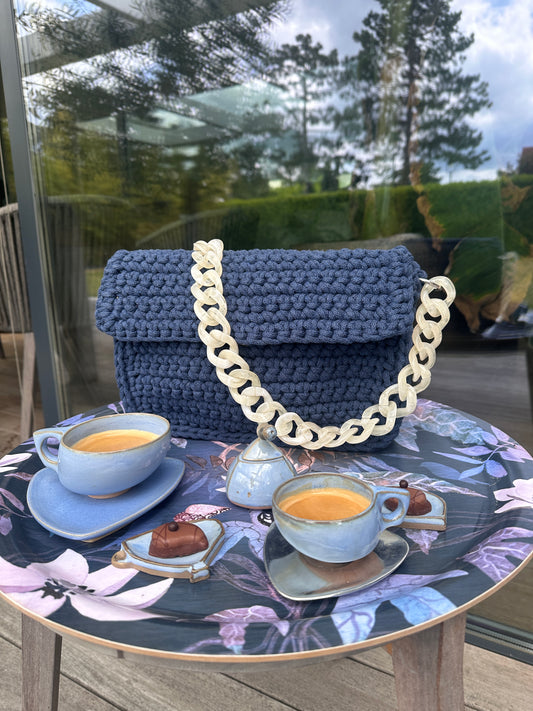 Eine geschlossene, gehäkelte Umhängetasche in einem dunklen Blauton, die auf einem Tisch liegt, mit einem auffälligen, cremefarbenen Kettenträger. Im Vordergrund stehen zwei blaue Tassen mit Espresso, neben einer passenden Kaffeekanne und einem kleinen Schokoladenstück, alles arrangiert auf einer Tischdecke mit floralen Mustern. Die Szene wirkt einladend und entspannt, möglicherweise ein Nachmittagskaffee im Freien.