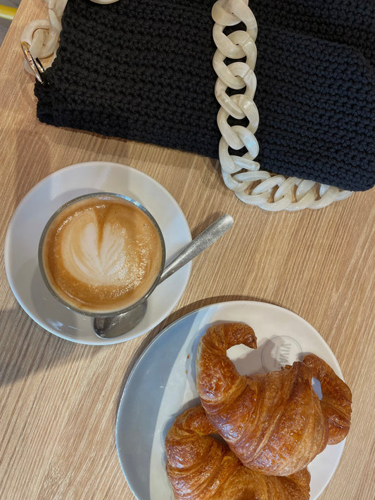 Ein gemütliches Kaffeearrangement mit einem Macchiato, der in einer weißen Tasse serviert wird und auf dessen Oberfläche ein Herzdesign aus Milchschaum zu sehen ist. Neben der Tasse befindet sich ein goldbrauner Croissant auf einem kleinen weißen Teller. Ein auffälliger, handgemachter Häkelbeutel mit schwarzen und weißen Elementen liegt teilweise im Bild.