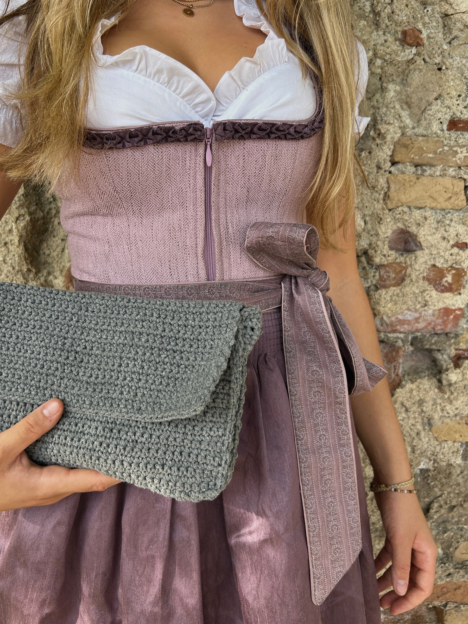 Ein stilvolles Foto einer jungen Frau in traditioneller Kleidung, die eine handgefertigte Häkeltasche hält. Sie trägt ein klassisches Dirndl in Rosa und Lila mit einer weißen Bluse und einer dekorativen Borte am Oberteil. Das Outfit wird durch eine farblich passende Schürze ergänzt. Die Kombination aus modernen und traditionellen Elementen vor der texturreichen Steinmauer bietet einen interessanten visuellen Kontrast.