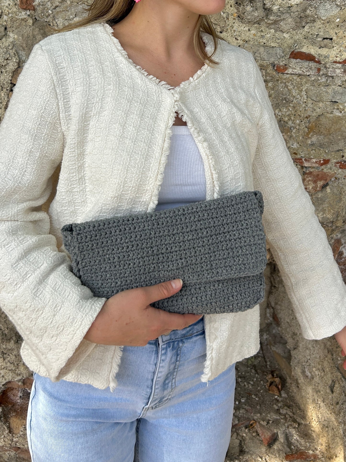 Ein Foto zeigt eine junge Frau, die einen schlichten, cremefarbenen Cardigan und eine weiße Bluse trägt, dazu eine klassische blaue Jeans. Sie hält eine stilvolle, handgemachte Häkeltasche in Grau. Die Tasche und die Kleidung stehen im Kontrast zur rauen Textur einer alten Steinmauer im Hintergrund.