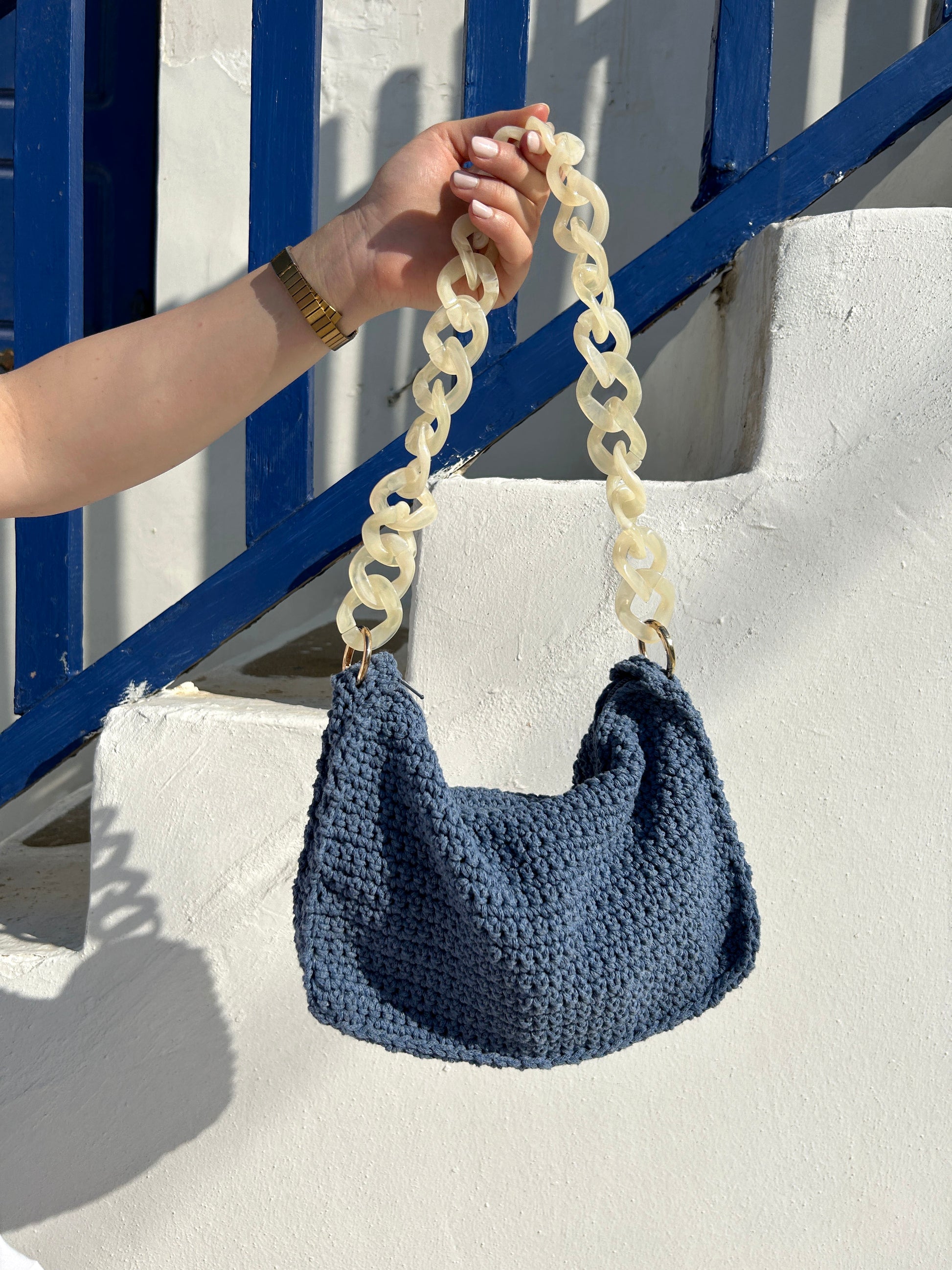 Ein stilvolles Foto einer handgefertigten Häkeltasche in Blau, die an einer hellen Wand neben einem blauen Holzgeländer aufgehängt ist. Die Tasche hat einen markanten, cremefarbenen Kettengurt, der gut zu dem lebendigen Blau der Tasche kontrastiert. Das leuchtende Sonnenlicht wirft Schatten und hebt die strukturierte Oberfläche der Tasche sowie die Details der Umgebung hervor, was das Bild visuell ansprechend macht.