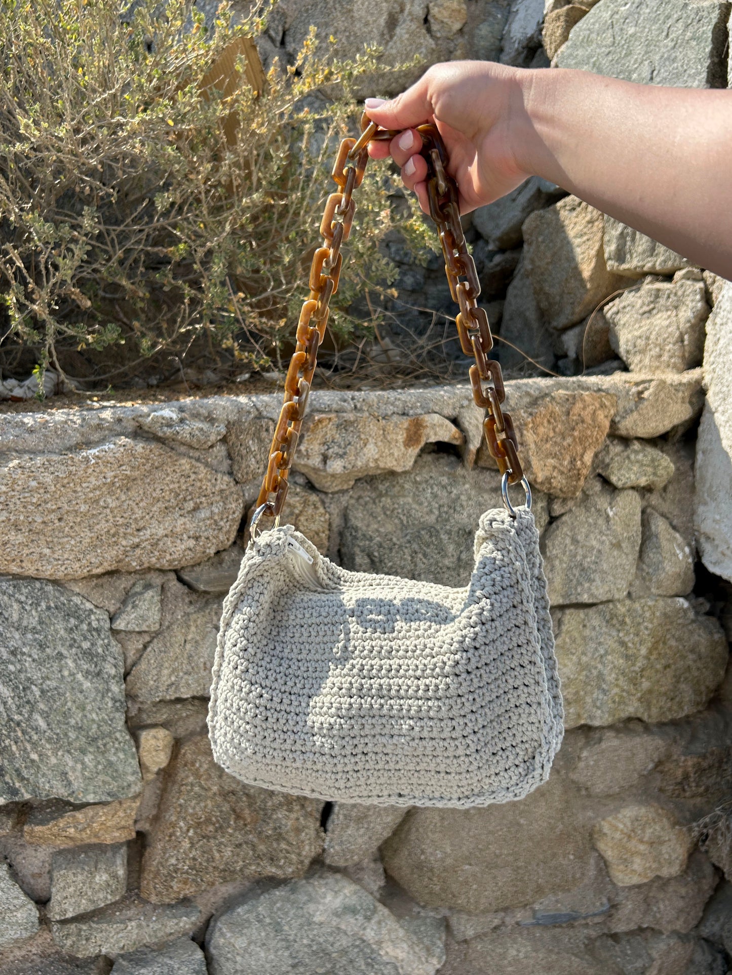 Das Foto zeigt eine sorgfältig gehäkelte Tasche in einem sanften Beigeton, die an einem robusten, dunkelbraunen Kettenriemen aufgehängt ist. Der Hintergrund aus rauen Steinblöcken ergänzt die weiche Textur der Tasche und bildet einen schönen Kontrast. Die natürlichen Farben der Tasche und der Umgebung verschmelzen harmonisch miteinander, während die robuste Struktur des Kettenriemens einen interessanten visuellen Akzent setzt.