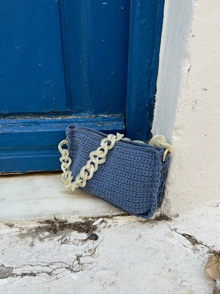 Ein charmantes Bild einer handgemachten Häkeltasche in Blau, die lässig an einer abgenutzten blauen Tür lehnt. Die Textur der Tasche kontrastiert schön mit der rauen, abblätternden Farbe der Tür und dem weiß verputzten Rahmen. Der grobe, weiße Kettengurt der Tasche fügt eine zusätzliche Texturschicht hinzu, die das handwerkliche Gefühl des Bildes verstärkt.