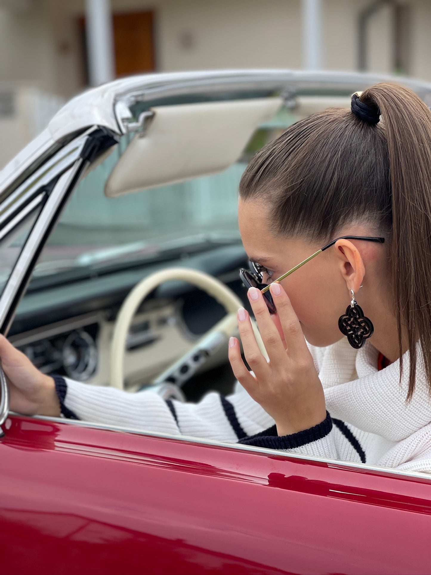 Das Bild zeigt eine junge Frau in einer weißen Strickjacke mit dunkelblauen Streifen, die sich an einem klassischen Cabriolet abstützt. Ihr Haar ist zu einem hohen Pferdeschwanz gebunden, und sie trägt große, dunkle Ohrringe im filigranen Design. Sie passt ihre Sonnenbrille an, während sie nachdenklich zur Seite blickt, was einen stilvollen und nachdenklichen Eindruck hinterlässt.