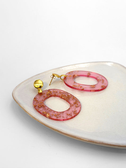 Ein Bild zeigt zwei kreisförmige Ohrringe aus rosa Resin, eingebettet mit Goldflocken, die auf einer keramischen Platte liegen. Die Ohrringe haben kugelförmige Stecker aus Messing (Brass).